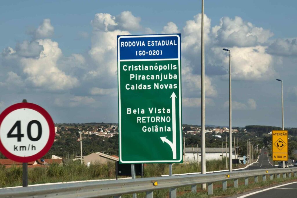 Foto de placas de sinalização da rodovia sentido Caldas Novas. Indicação da velocidade permitida, retorno no sentido de Goiânia e Bela Vista e outra placa de atenção à rotatória a 100 metros.