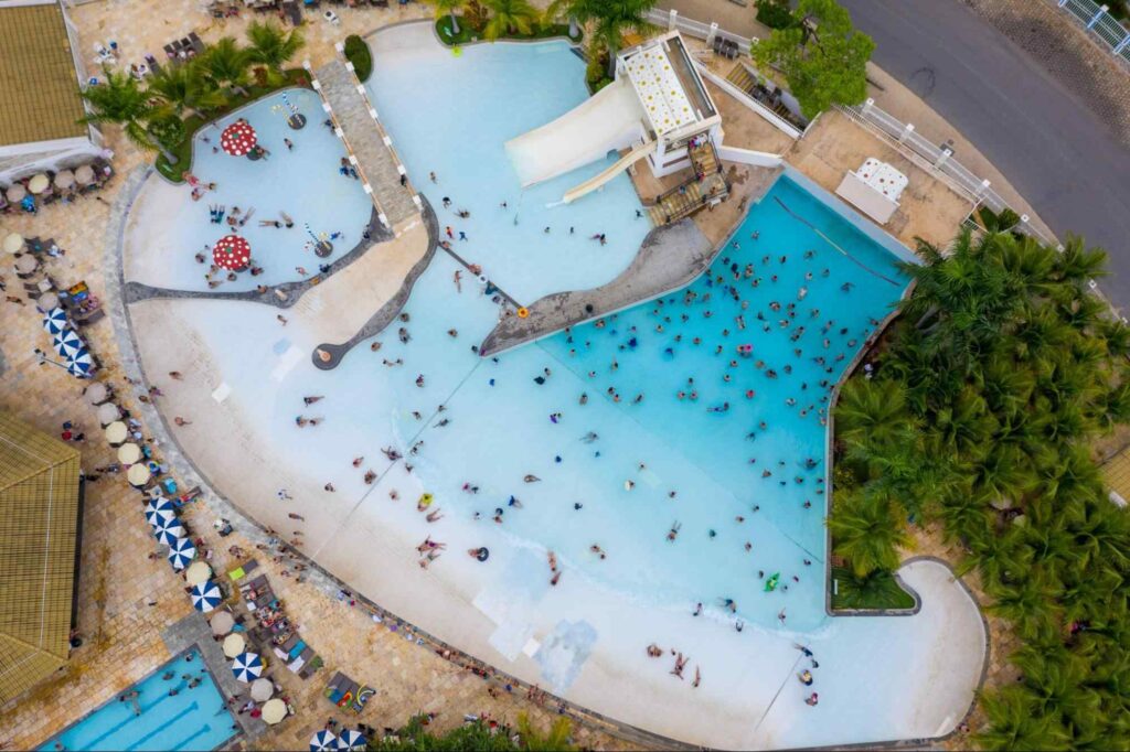 Vista de cima de uma das grandes piscinas do Water Park em Caldas Novas GO. Diversas pessoas na água, piscina cercada de árvores, guarda-sóis e espreguiçadeiras.