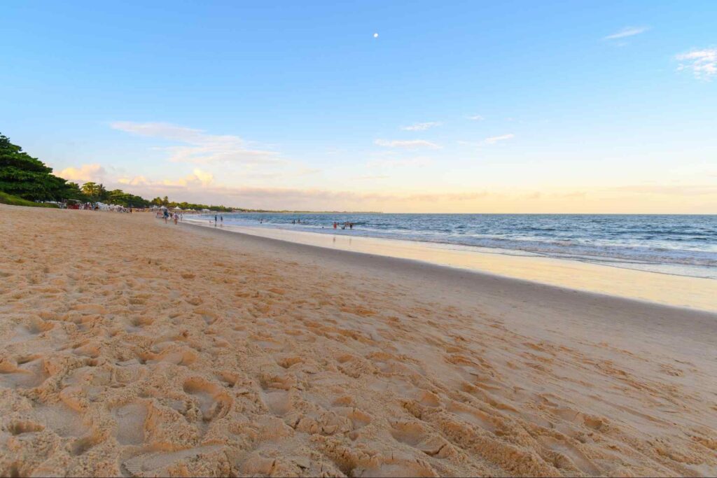 Faixa de areia dourada da Praia de Taperapuan, Porto Seguro. O mar está tranquilo e recuado. Alguns banhistas caminham pela praia e entram na água.