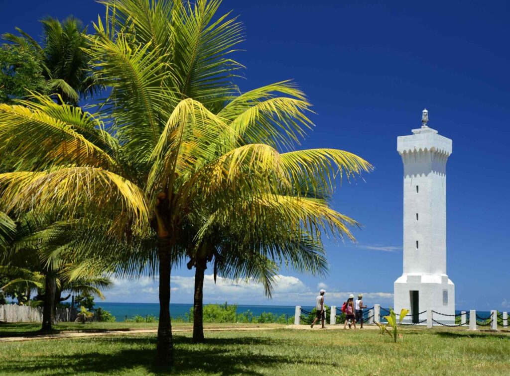 Farol de Porto Seguro, pequena torre solitária pintada de branco e situada em um gramado. Ao fundo, aparece o mar.