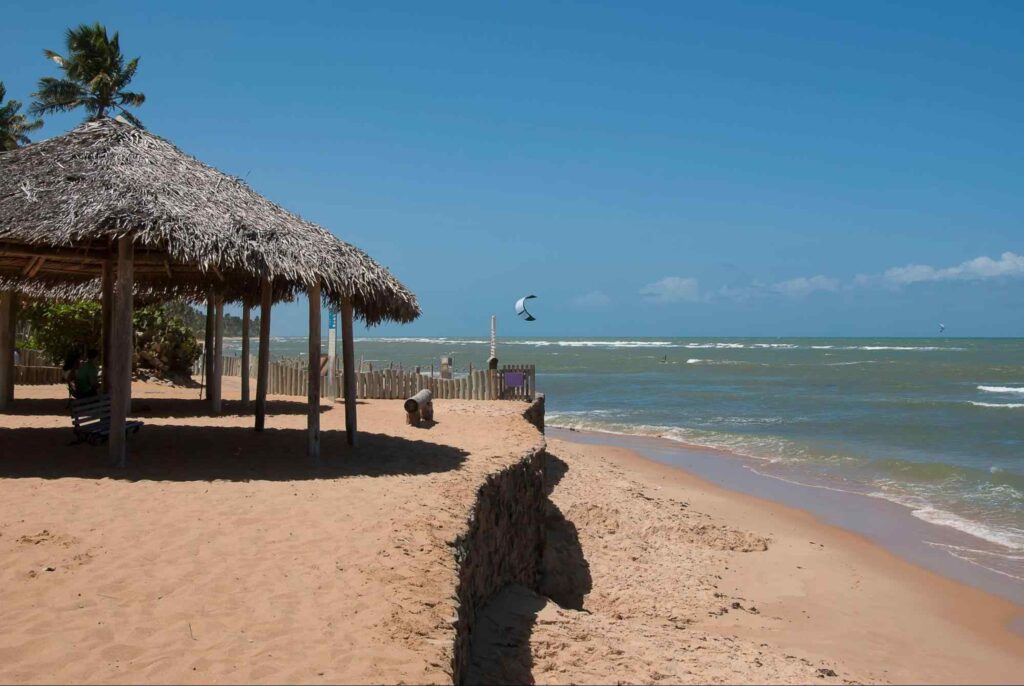 Foto da Praia do Forte, em Mata de São João. Bangalô sem paredes ao lado esquerdo da imagem, fazendo sombra em parte da areia em frente ao mar, ao lado direito.