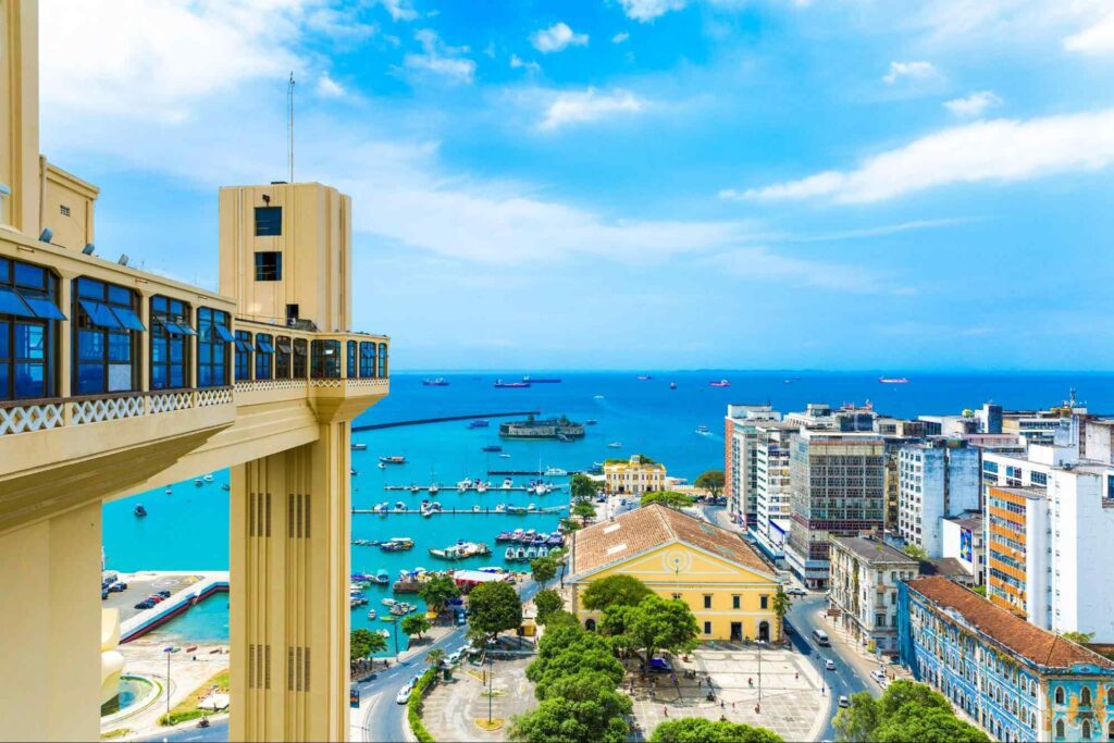 Foto do Elevador Lacerda (Salvador BA). Elevador ao lado esquerdo da imagem, com vista para o Mercado Municipal de Salvador abaixo, ao lado dos prédios da Cidade Baixa, e o mar repleto de barcos e navios.