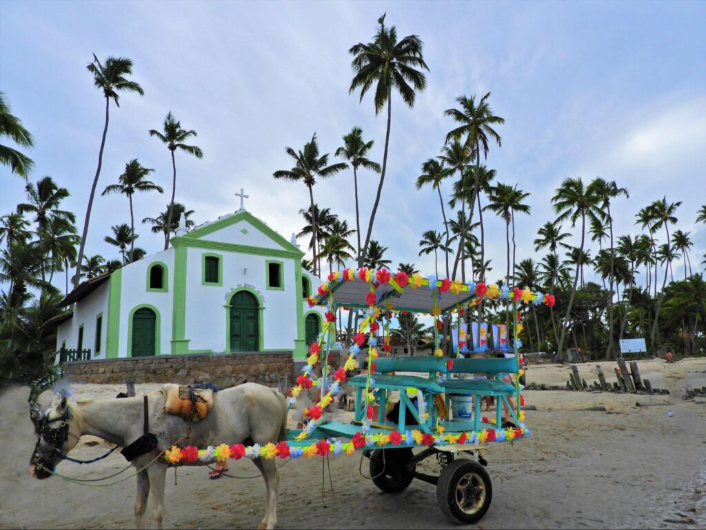 Igreja da Praia dos Carneiros, em Tamandaré. A capela de fachada branca e verde está rodeada pela areia da praia e por coqueiros. À sua frente, está parada uma charrete rústica decorada com flores coloridas.