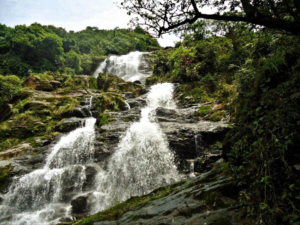 Cachoeira da fumaça vista de baixo. A queda d’água tem dois estágios entre as rochas, antes de chegar ao final. As pedras são cobertas de limo e existe vegetação dos dois lados.