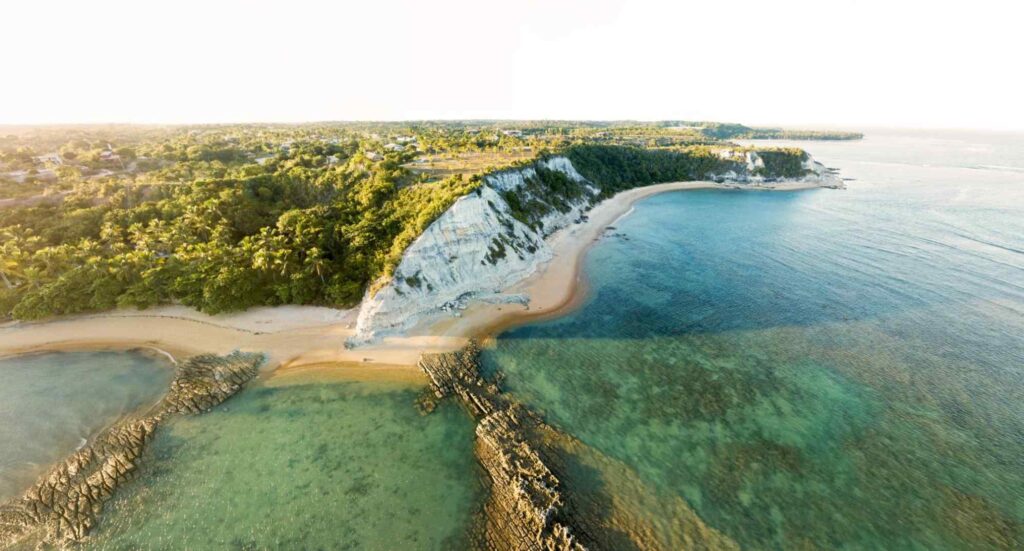 Vista aérea panorâmica da Praia do Espelho. Falésias altas cobertas pela vegetação, com faixa de areia em frente ao mar cristalino com recifes fora d’água.