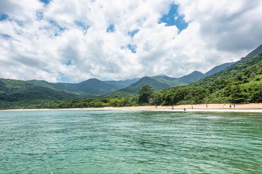 Mar esverdeado da Praia da Parnaioca. Ao fundo, aparecem alguns banhistas, que caminham pela estreita faixa de areia clara limitada pelos montes cobertos de árvores.