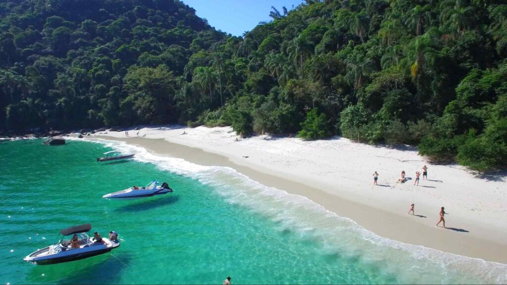 Praia do Dentista. Poucas pessoas caminham pela faixa de areia branca, que é cercada por uma mata densa e verde. O mar cristalino tem algumas pequenas embarcações.