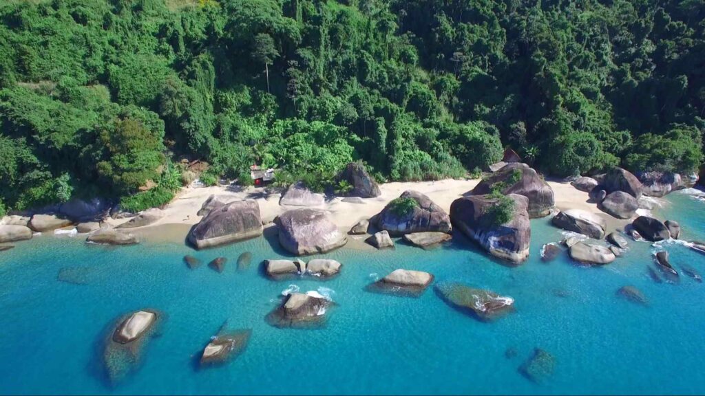 Vista aérea da Praia Secreta, Angra dos Reis. O mar de cor azul vibrante tem muitas rochas grandes e cinzas. Depois da pequena faixa de areia branca, há uma floresta densa com árvores altas.
