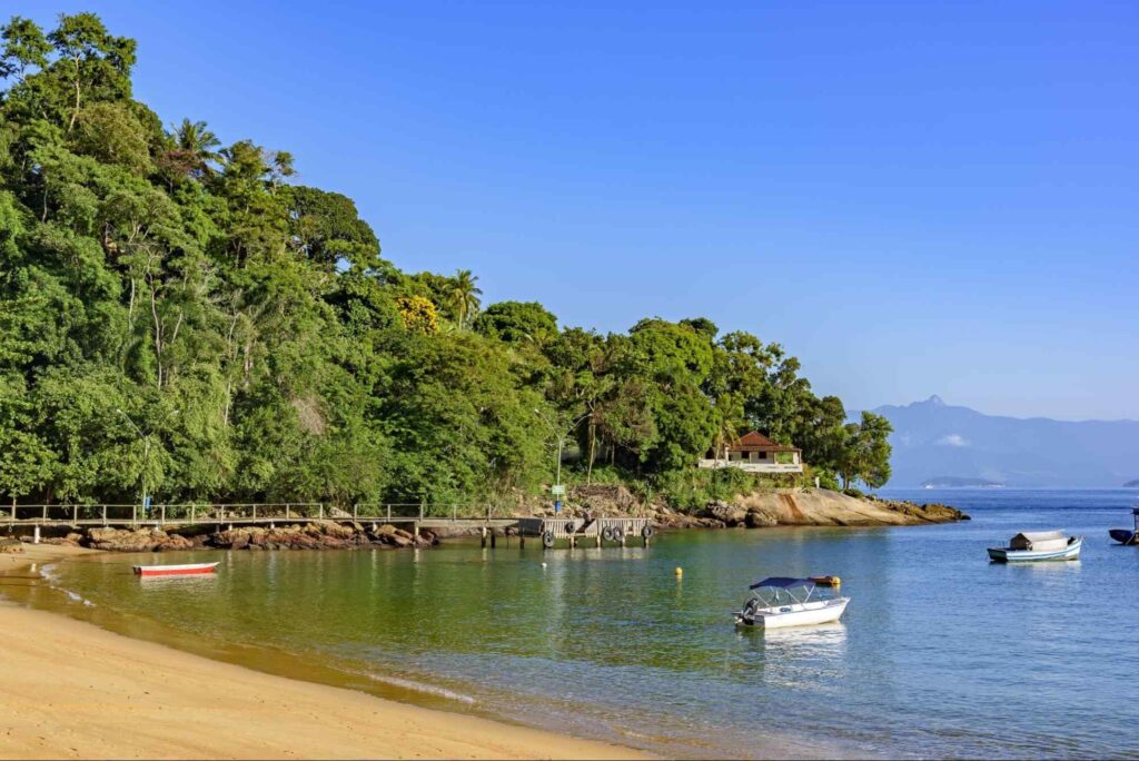 Final da Praia Vermelha da Ilha Grande. A faixa de areia dourada termina em uma ponta coberta por floresta nativa. Alguns barquinhos flutuam na água de tonalidade verde profunda.
