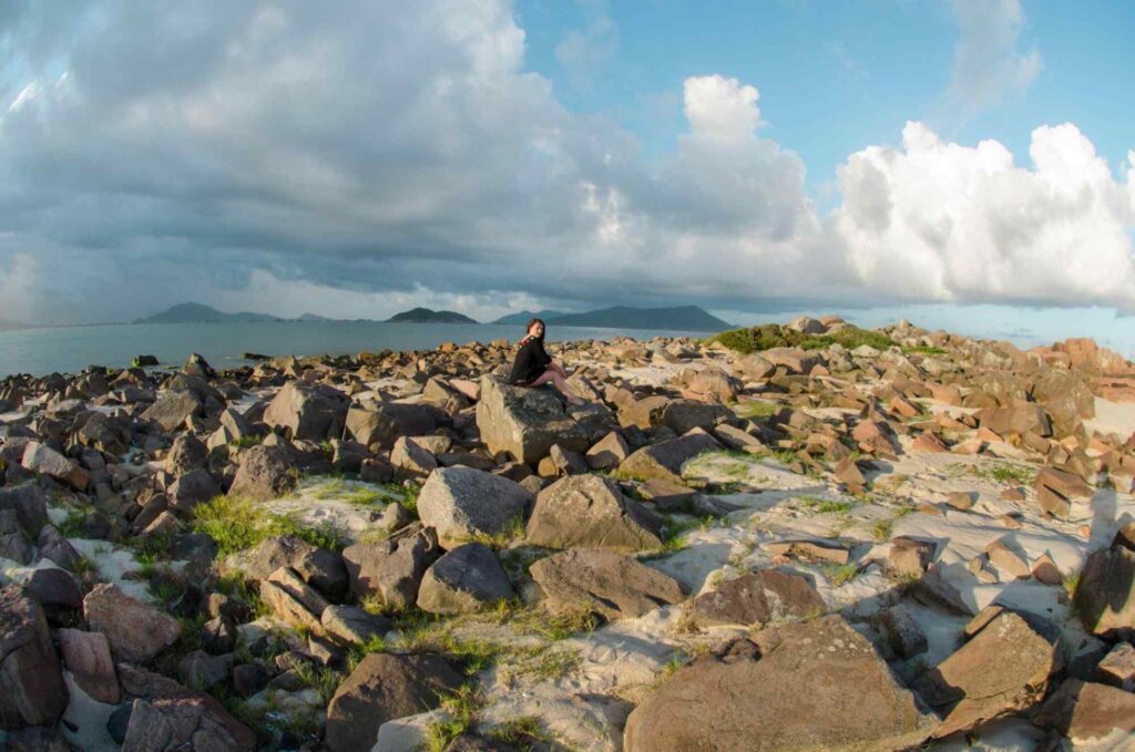 Foto de mulher sentada nas formações rochosas da Praia da Pinheira em dia ensolarado, com grandes nuvens no céu.