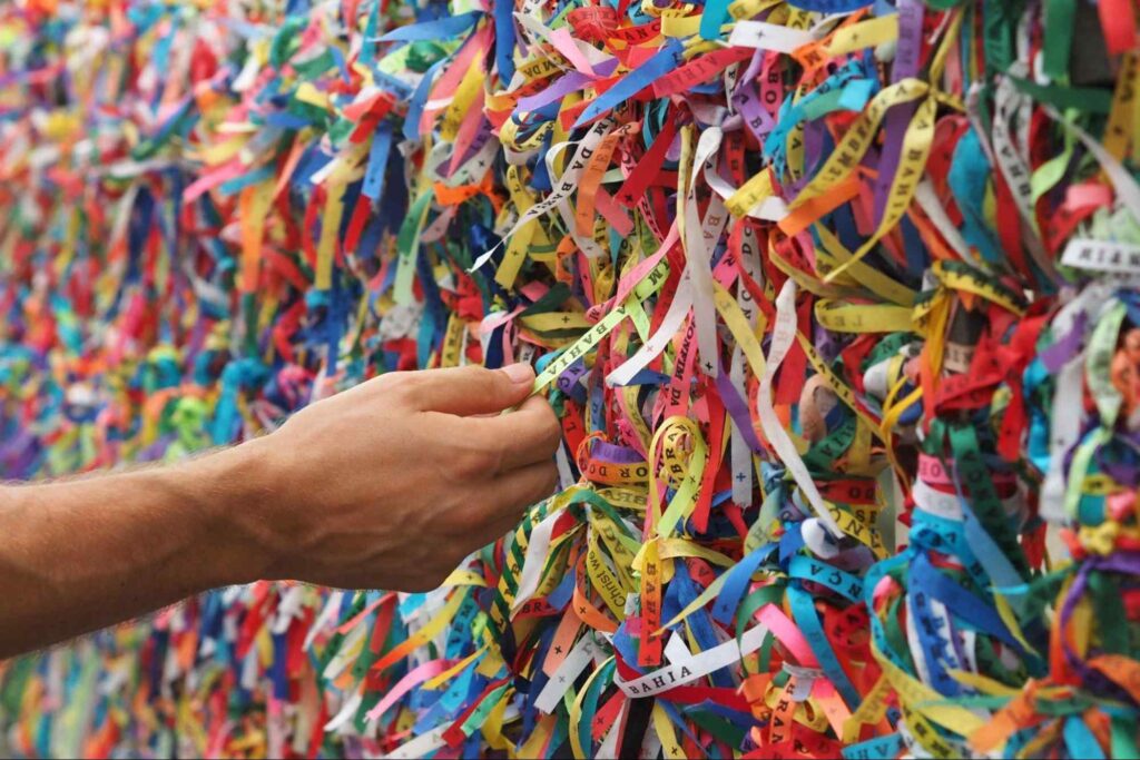 Homem tocando uma fita do Senhor do Bonfim. Há várias fitas de cores diferentes amarradas a uma grade.