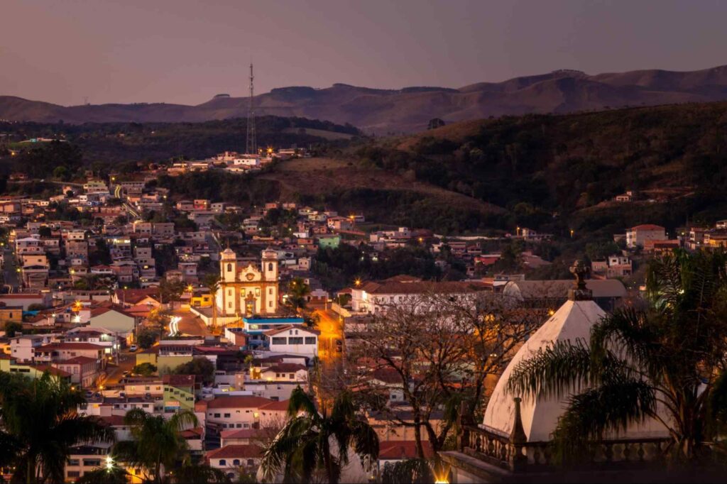 Vista da cidade de Congonhas ao anoitecer. Construções iluminadas por lâmpadas amareladas estão rodeadas pelas serras verdes e o céu está escurecendo