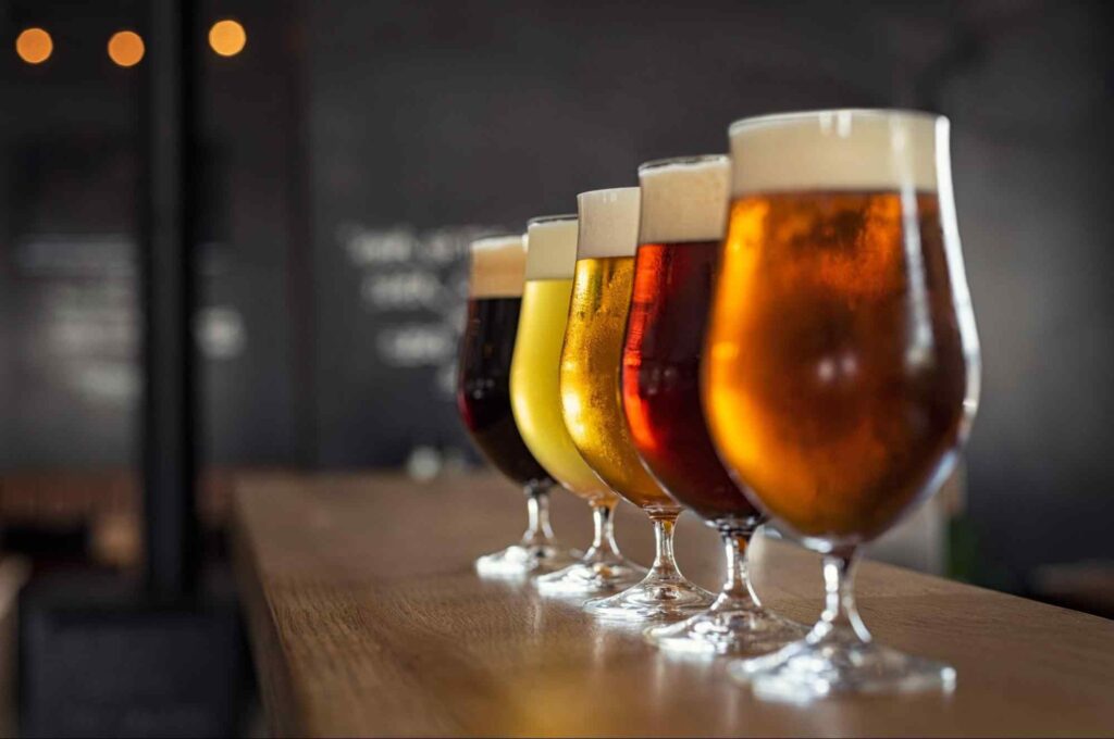 Cinco copos de cerveja enfileirados sobre um balcão de madeira. Eles contêm cervejas de colorações diferentes e parecem estar gelados.