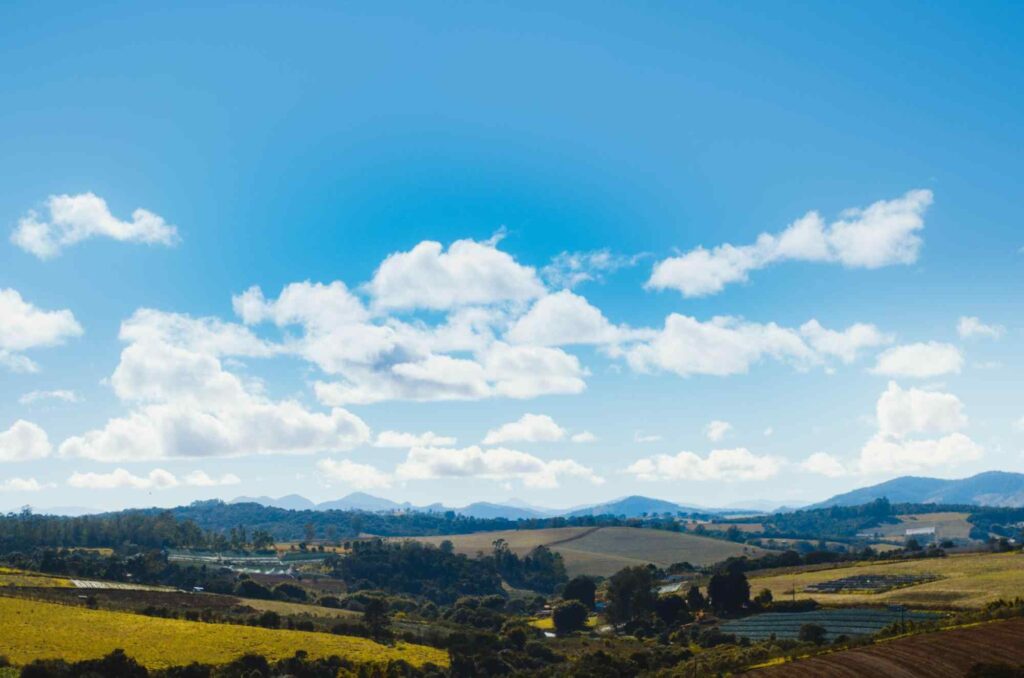 Paisagem com campos verdes, árvores, morros e serra ao fundo, com céu azul e nuvens