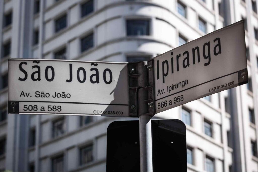 Duas placas de sinalização urbana indicando nomes de ruas perpendiculares. Uma delas tem os dizeres “São João - Avenida São João”. A outra diz “Ipiranga - Avenida Ipiranga -”