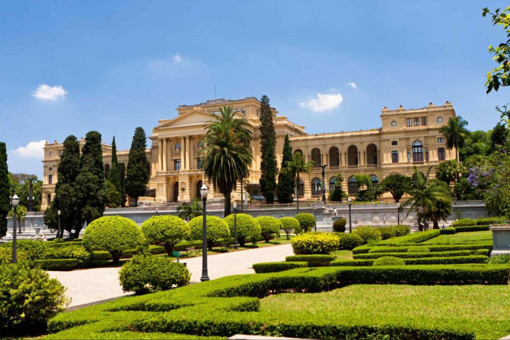 Museu do Ipiranga, que é um antigo palácio de arquitetura neoclássica. A fachada amarela ornamentada tem colunas e arcos. Ao redor da construção, canteiros bem cuidados, com gramados, arbustos e árvores decorativas.