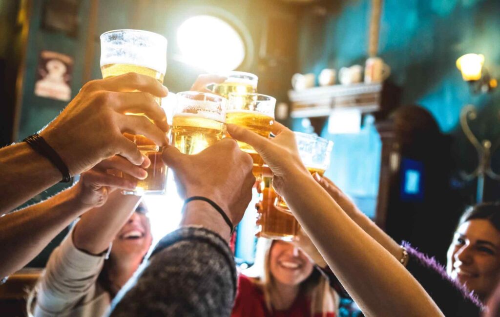 Grupo de sete pessoas brindando com copos de cerveja. É possível ver as mãos, partes dos antebraços e dos rostos de algumas delas.