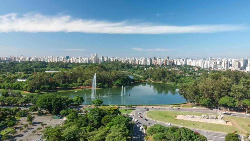 Vista aérea do Parque Ibirapuera, mostra um grande lago com fontes que jogam água para o alto. Ao redor, muitas árvores que cobrem de verde a maior parte da paisagem.