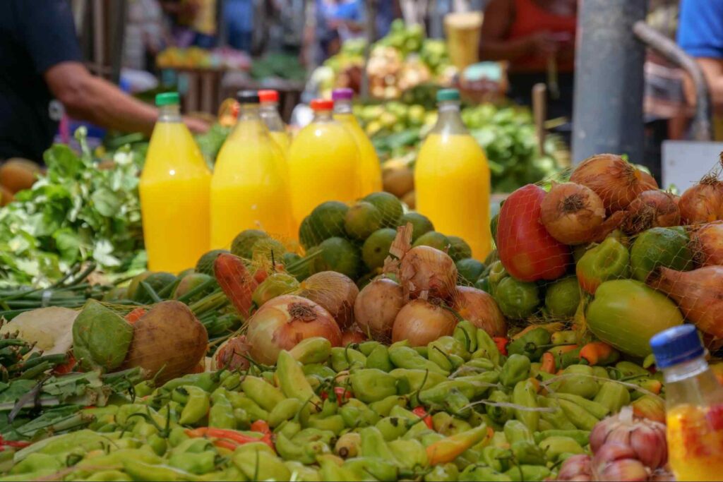 Produtos expostos no Ver-o-Peso. Há frutas e legumes diversos, como cebolas, pimentões e bananas, exibidos sobre uma mesa. Entre eles, algumas garrafas de tucupi.
