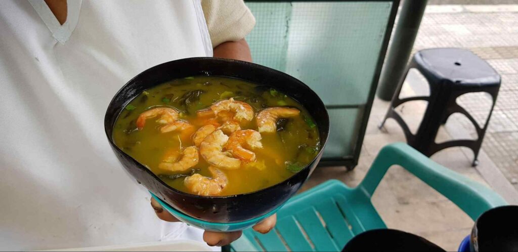 Mão de um homem segurando uma tigela cheia de tacacá, prato típico do Pará. É uma sopa composta por um caldo amarelo espesso, ervas e temperos verdes e grandes camarões.