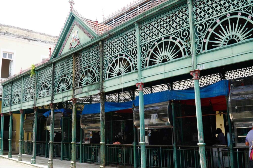 Fachada do Mercado Ver-o-Peso. Estrutura de ferro esverdeado, com colunas e detalhes decorativos no estilo art nouveau. No interior estão algumas pessoas e barracas de feira