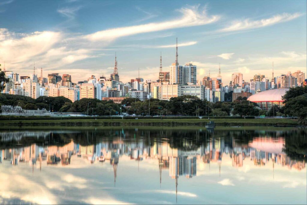 Paisagem do Parque Ibirapuera, com área verde e prédios ao redor refletidos na água de um dos lagos do local.