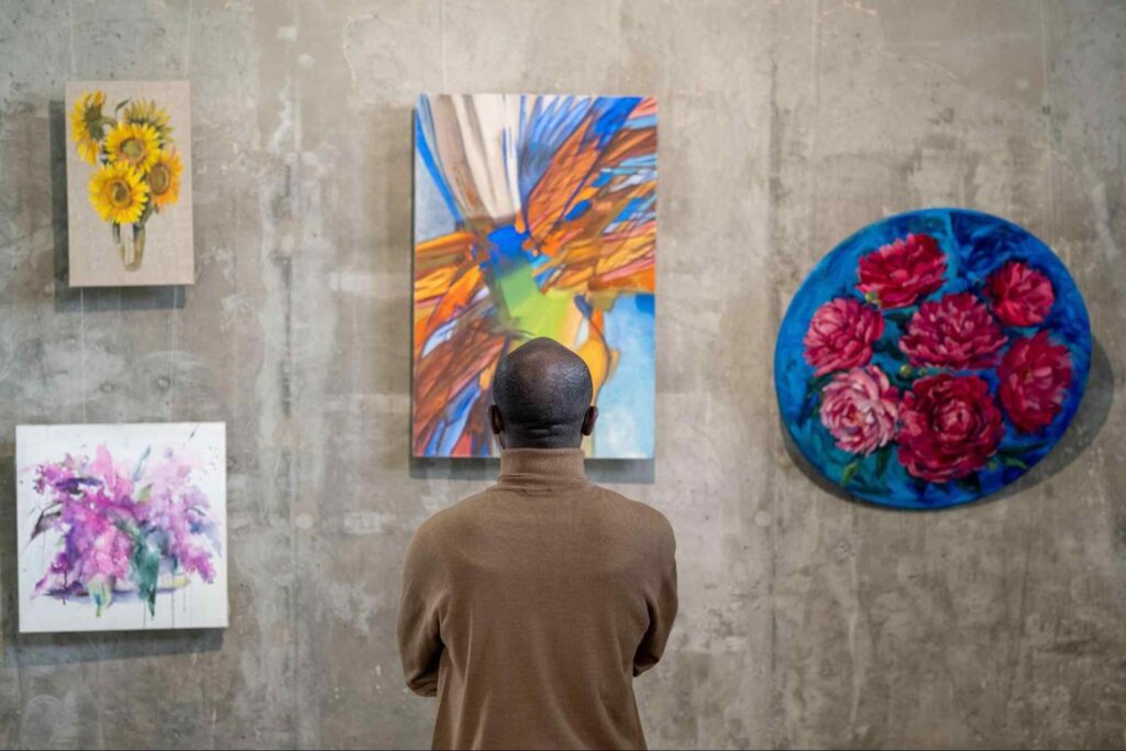 De costas para a foto, um homem examina quatro pinturas penduradas em uma parede de concreto. As peças têm traços contemporâneos e trazem motivos florais e abstratos.