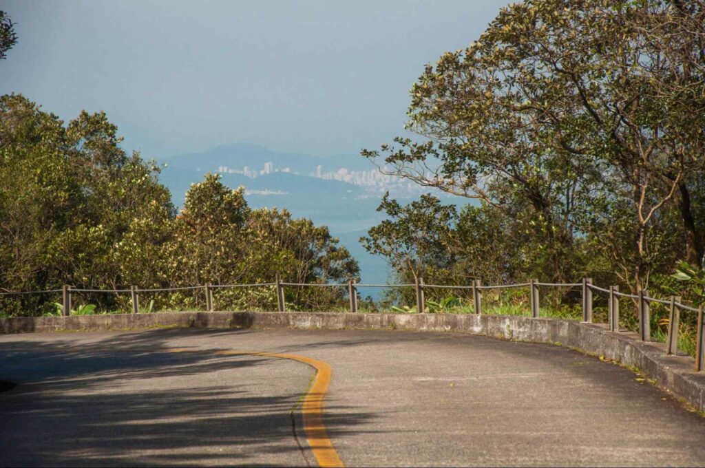 Trecho da SP-148, conhecida como Estrada Caminho do Mar, que liga o litoral de São Paulo ao planalto paulista. Além do fragmento da estrada, é possível ver parte das serras e as silhuetas dos prédios de uma cidade.