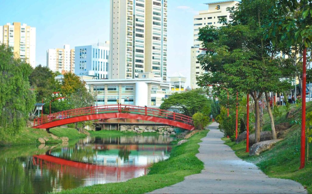 Lago sobre o qual passa uma pequena ponte vermelha em estilo japonês. Nos arredores, há uma pista de caminhada com luminárias orientais e algumas árvores. Ao fundo, vê-se os prédios de fachada clara da cidade.