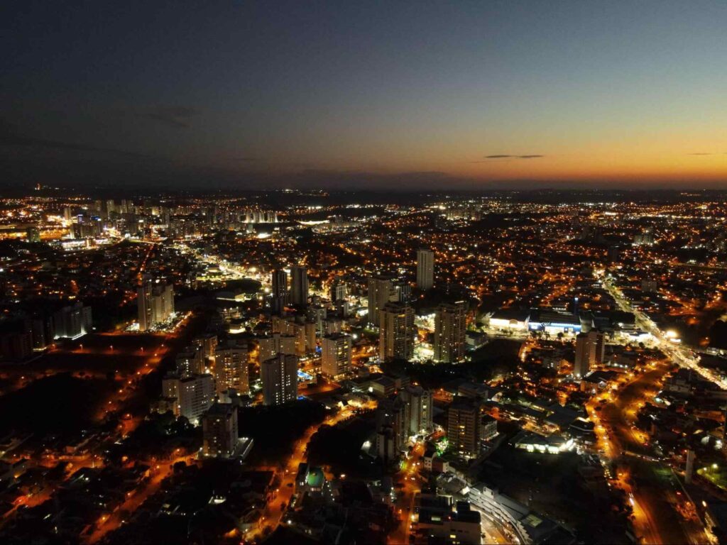 Vista aérea noturna de Sorocaba. As vias e as construções estão iluminadas por pontos de luz amarela e azul. O céu ainda está um pouco alaranjado por causa do pôr do sol