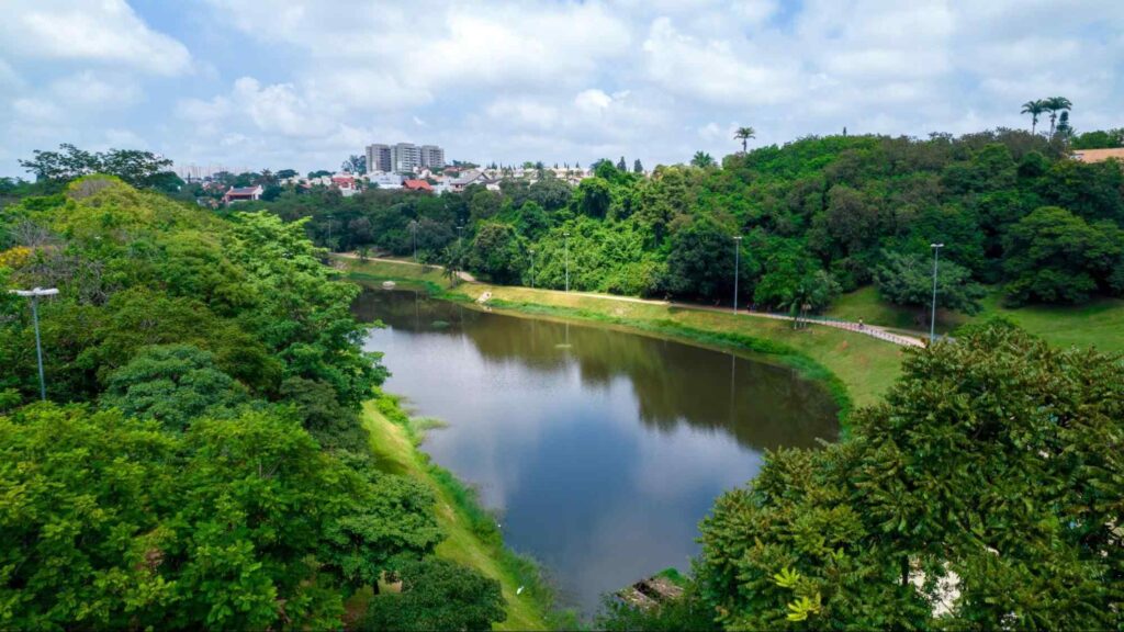 Lago de águas escuras no Parque do Campolim. Ao redor estão áreas arborizadas e trechos de gramado aberto. Há ainda alguns postes de luz. Ao fundo, é possível ver algumas construções da cidade.