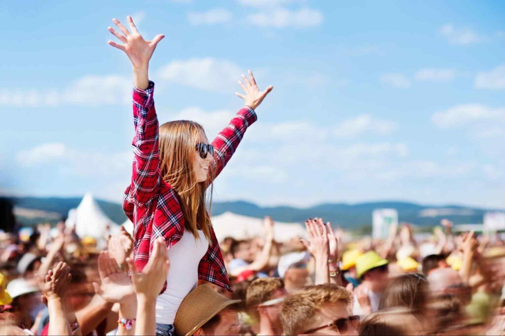 Foto de público em show ao ar livre. A foto foca em uma jovem em cima dos ombros de um homem, enquanto joga seus braços para cima.