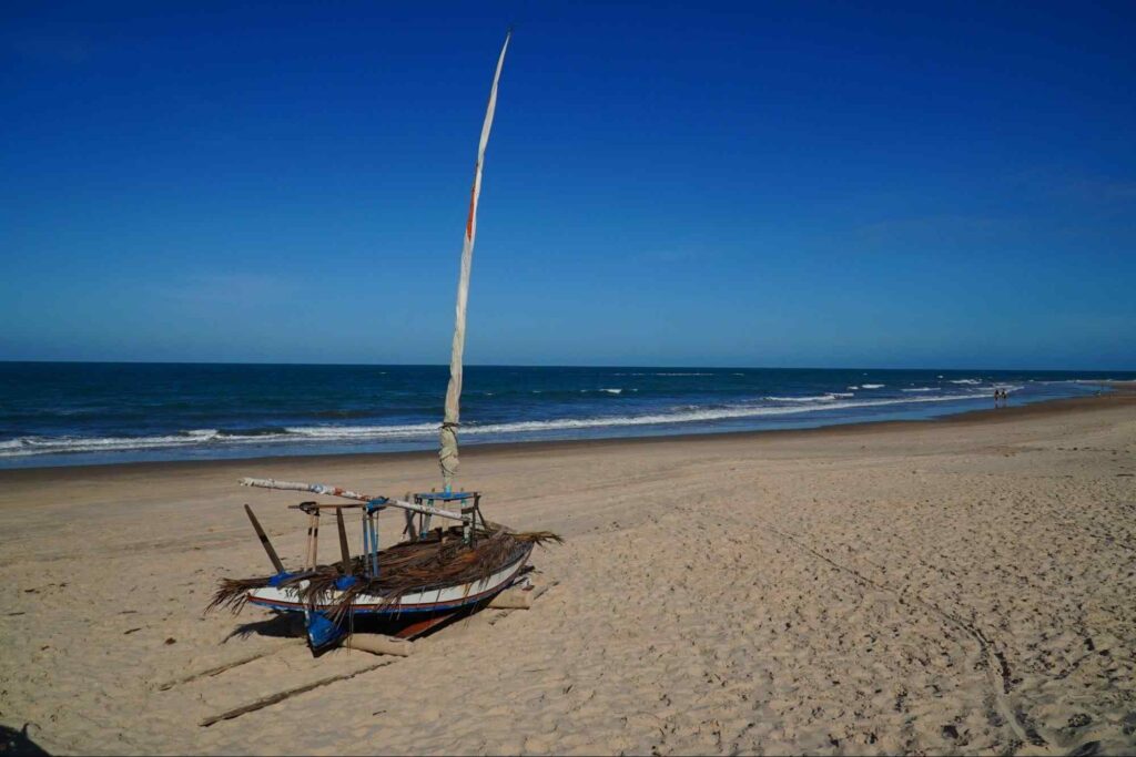 Pequena jangada parada na areia da praia. A faixa de areia clara está deserta e se estende até um mar azul e com poucas ondas. O céu está aberto