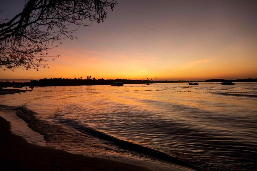 Mar da Ilha de Boipeba durante o pôr do sol. A água forma ondas discretas e está ofuscada pelo céu. É possível ver as silhuetas das árvores da vegetação nativa e de duas pequenas embarcações.