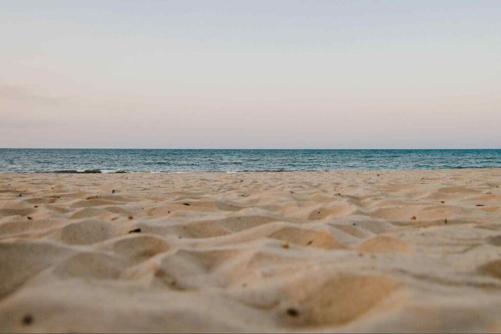 Foto tirada do ponto de vista do chão na areia em frente ao mar. Céu com um tom levemente acinzentado em contraste com o azul do mar.