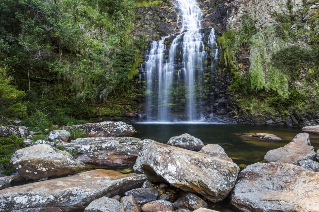 Ao fundo está a alta Cachoeira da Farofa, cercada por vegetação. O lago que recebe a queda d’água parece raso e tem tom esverdeado. Em primeiro plano estão algumas pedras.