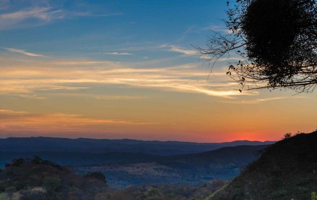 Pôr do Sol na Serra do Cipó. O horizonte tem tom alaranjado enquanto o resto do céu vai escurecendo. a vegetação do Parque é rasteira e repleta de pedra e imperfeições.