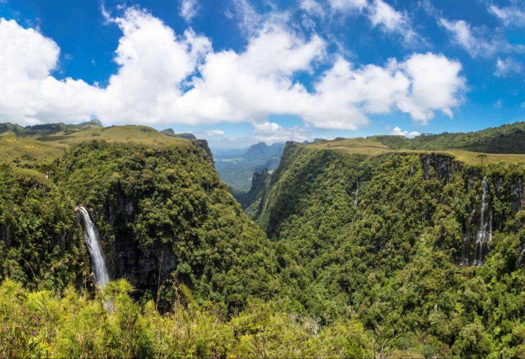 Vista do Cânion do Espraiado. Ele surge como uma grande fenda entre os chapadões densamente cobertos por árvores verdes. No canto esquerdo da foto, aparece uma queda d’água que flui em direção ao vale.