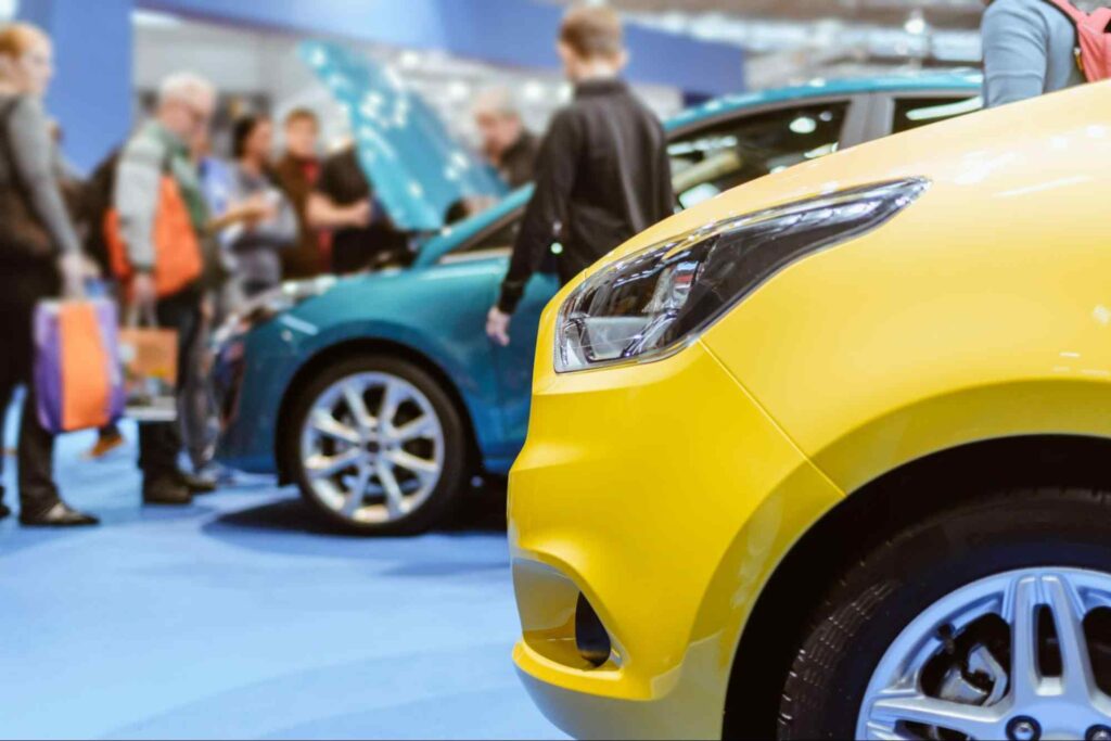 Foto de feira de carros esportivos com a frente de um carro amarelo tomando conta do lado direito da imagem. Ao lado esquerdo, imagem desfocada de carro azul com capô aberto e participantes observando seu motor.