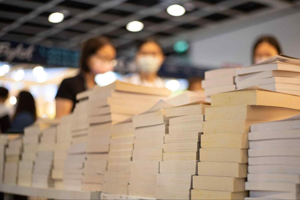 Livros empilhados em cima de uma mesa em uma feira literária. Em segundo plano, algumas pessoas parecem examinar os volumes.