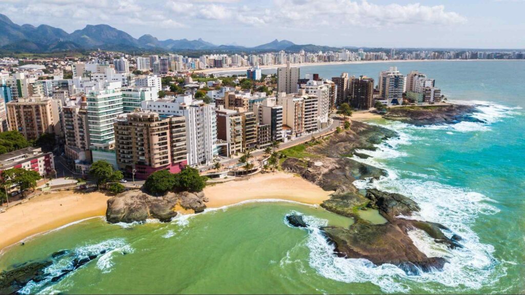Vista aérea da cidade de Guarapari ES. Contraste da paisagem com o mar do lado direito e a cidade à orla da praia. Algumas pedras se mesclam com a faixa de areia da praia.