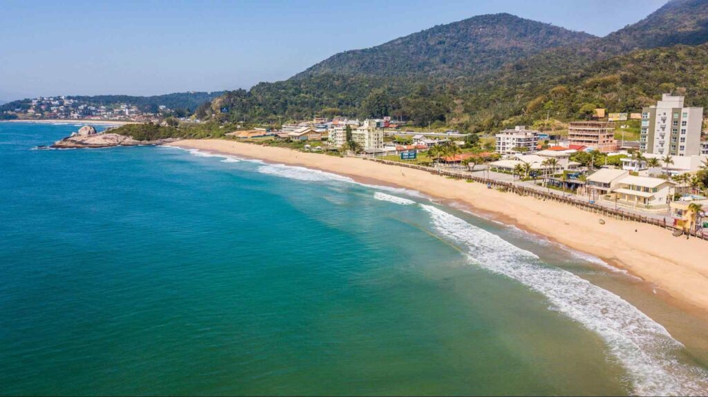 Imagem aérea da Praia da Ilhota, com o mar tomando conta de grande parte da foto. Faixa de areia extensa, com uma orla repleta de vegetação, casas e prédios.
