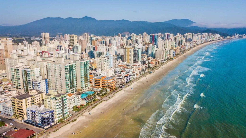 Foto aérea da Meia Praia, Itapema. Diversos prédios com diferentes alturas em frente à orla da praia, com faixa de areia clara que leva ao mar esverdeado