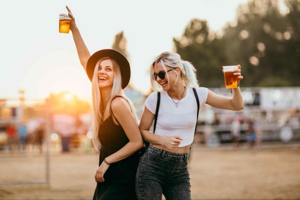 Duas mulheres com copos de cerveja nas mãos, dançando em festival ao ar livre. Luz do entardecer ao fundo, com árvores e quiosques borrados ao fundo da imagem. 