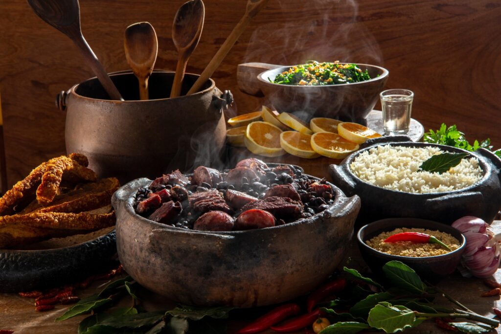Mesa disposta com várias panelas de ferro, recheadas de feijoada, arroz, laranja e outros acompanhamentos