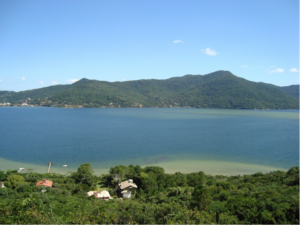 Lagoa da Conceição em Florianópolis com dunas, água azul e montanhas ao fundo sob céu ensolarado.
